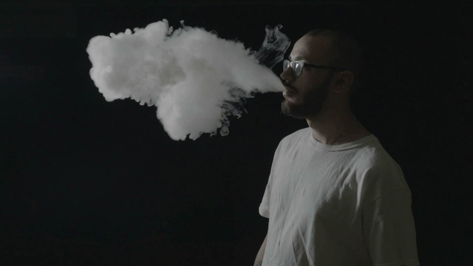 1920x1080 Young hipster boy with glasses vaping from a vaporizer and exhaling large  clouds of smoke repeatedly in a professional dark studio Stock Video  Footage