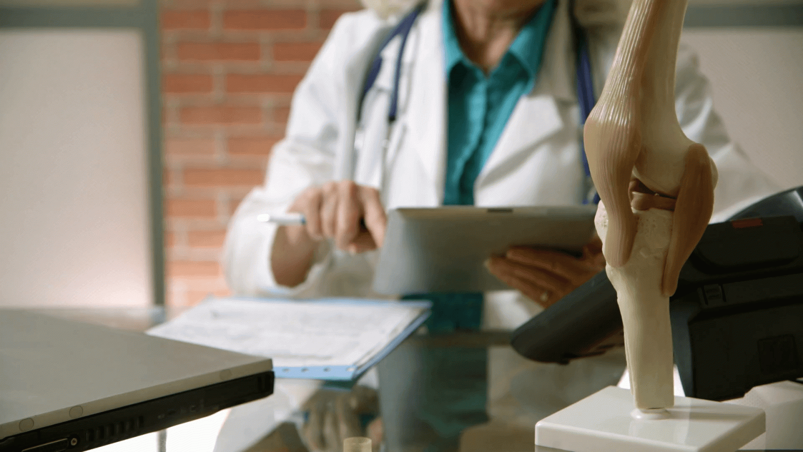 1920x1080 focus on knee model as orthopedic doctor read notes in background